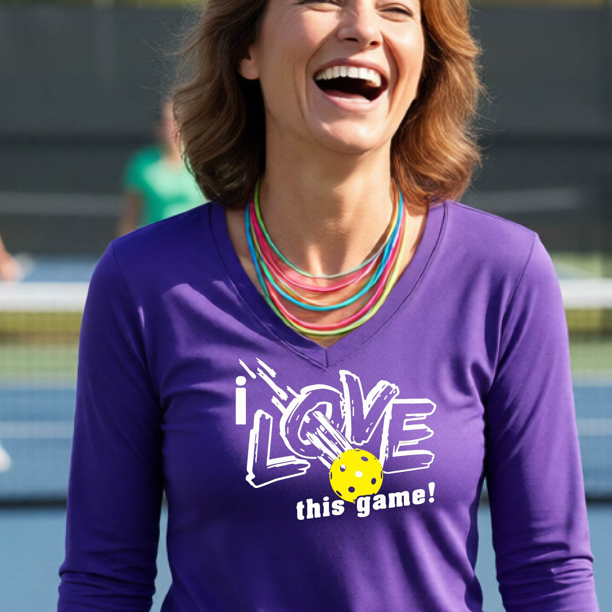 A woman wearing a purple long-sleeve V-neck athletic shirt with a graphic that reads 'i LOVE this game!' in white, with a yellow pickleball over the letter O. She is smiling and standing on a p ikleball court.