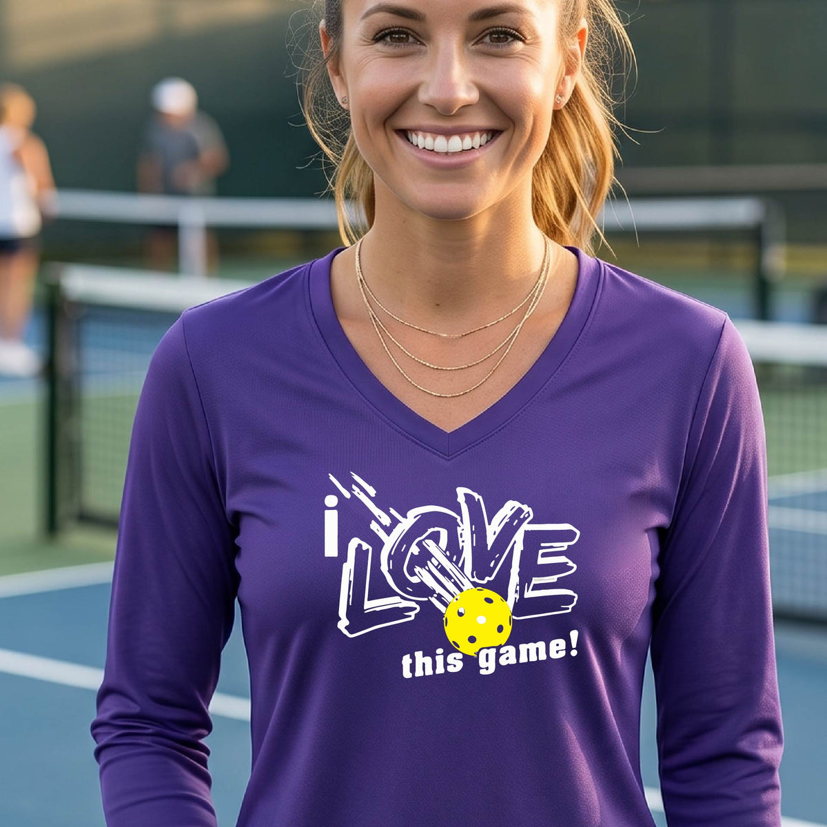 A woman on a pickleball court wearing a purple long-sleeve V-neck athletic shirt with a white graphic that reads 'i LOVE this game!' and a yellow tennis ball illustration.