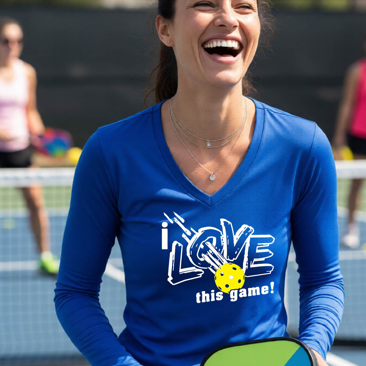 A woman on a pickleball court wearing a royal blue long-sleeve V-neck athletic shirt with a white graphic that reads 'i LOVE this game!' and a yellow tennis ball illustration.