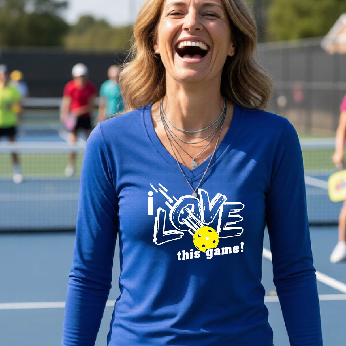 A woman wearing a royal blue long-sleeve V-neck athletic shirt with a graphic that reads 'i LOVE this game!' in white, with a yellow pickleball over the letter O. She is smiling and standing on a p ikleball court.