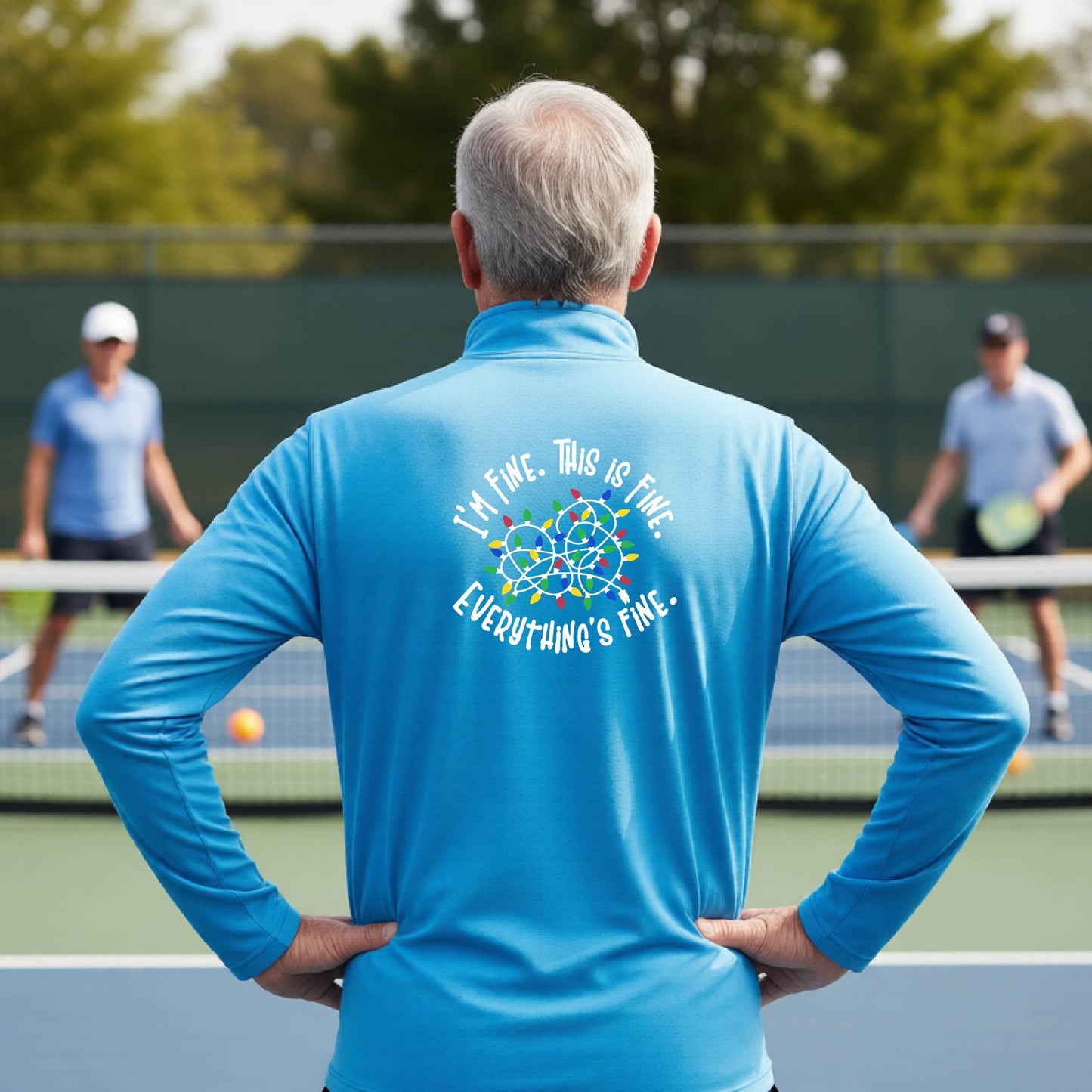 A man viewed from behind on an outdoor pickleball court, wearing a  bright light blue long-sleeve performance shirt with a colorful web graphic and the text 'I'M FINE, THIS IS FINE, EVERYTHING'S FINE.' printed on the back.