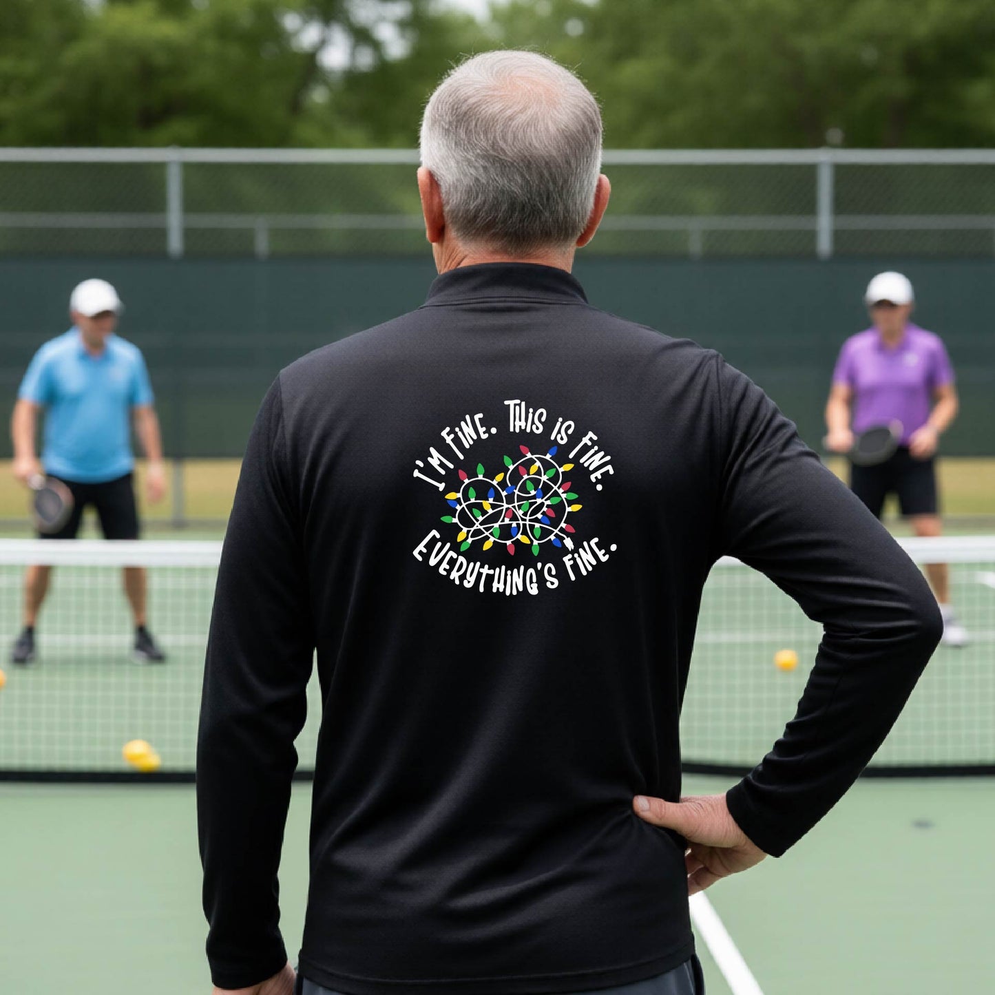 A man viewed from behind on an outdoor pickleball court, wearing a black long-sleeve performance shirt with a colorful web graphic and the text 'I'M FINE, THIS IS FINE, EVERYTHING'S FINE.' printed on the back.