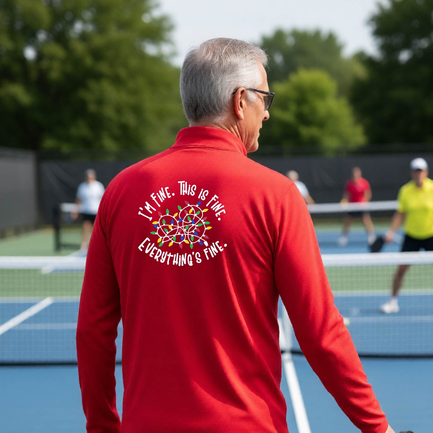 A man viewed from behind on an outdoor pickleball court, wearing a red long-sleeve performance shirt with a colorful web graphic and the text 'I'M FINE, THIS IS FINE, EVERYTHING'S FINE.' printed on the back.