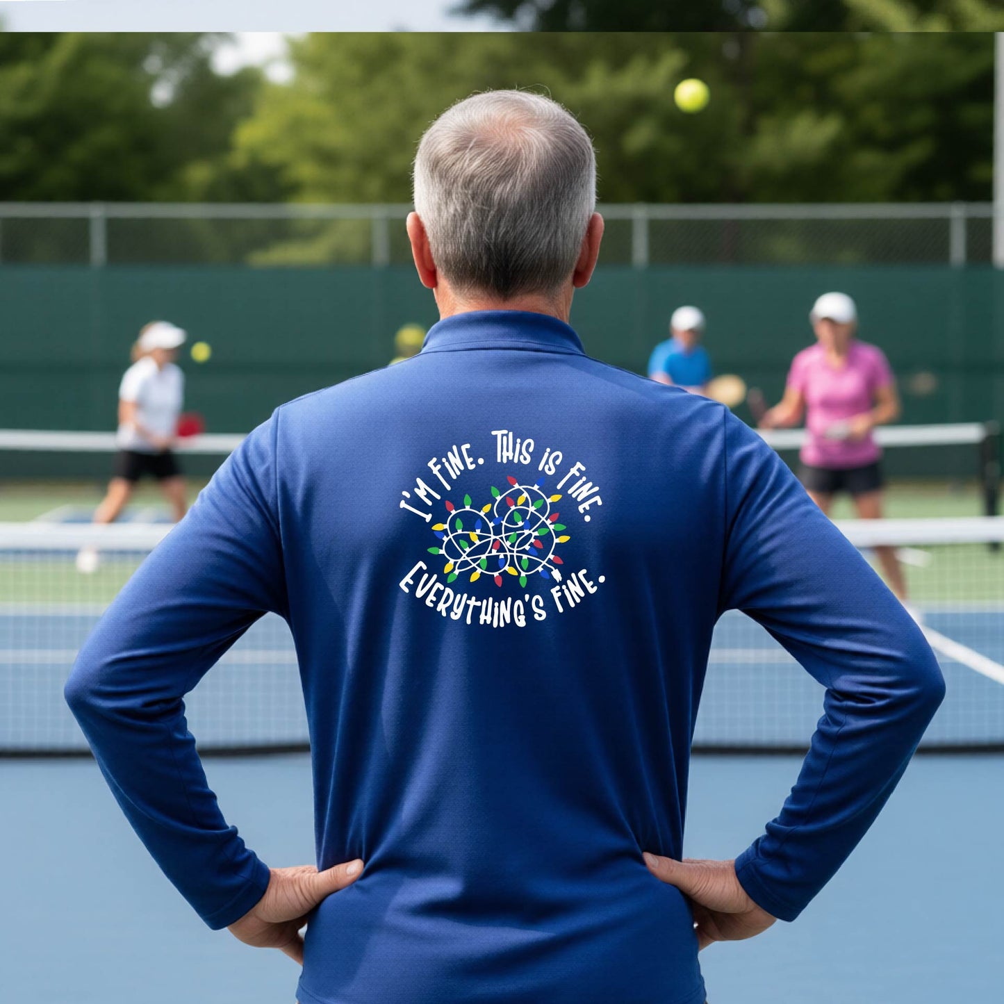A man viewed from behind on an outdoor pickleball court, wearing a royal blue long-sleeve performance shirt with a colorful web graphic and the text 'I'M FINE, THIS IS FINE, EVERYTHING'S FINE.' printed on the back.