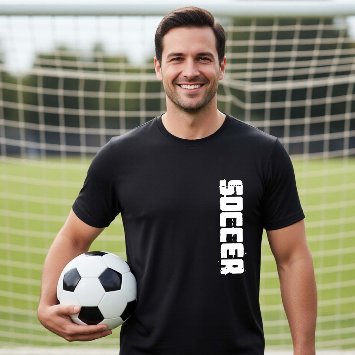 A smiling middle-aged man wearing a black athletic t-shirt with the word 'SOCCER' printed vertically in white on the right side of the chest, holding a black and white soccer ball in front of a soccer goal net.