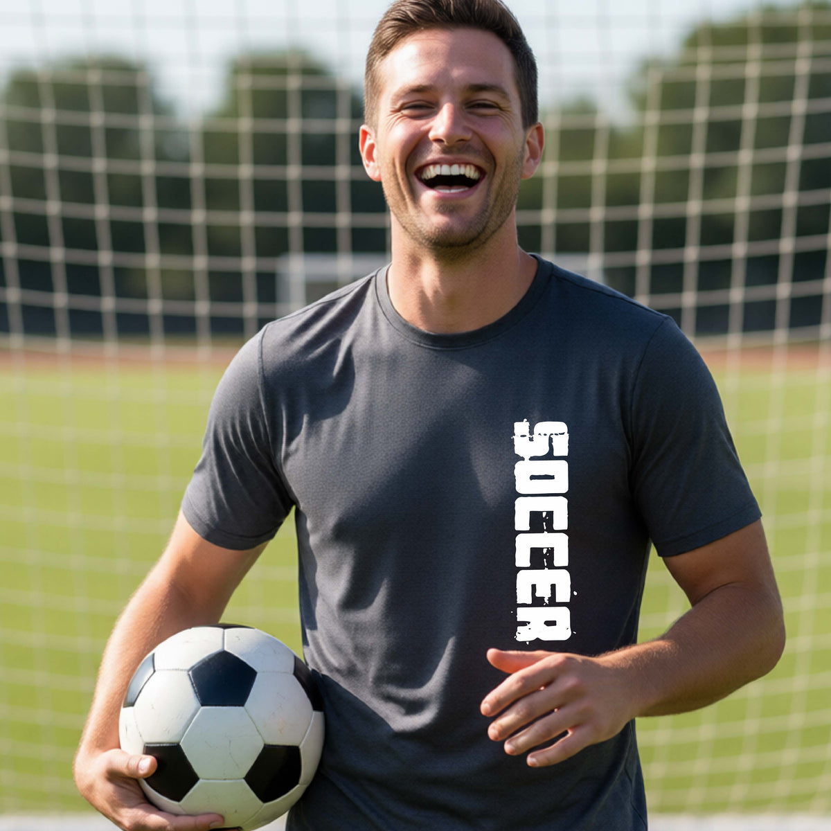 A smiling man on a soccer field holds a black-and-white soccer ball while wearing a dark gray athletic T-shirt with the word 'SOCCER' printed vertically in white on the front.