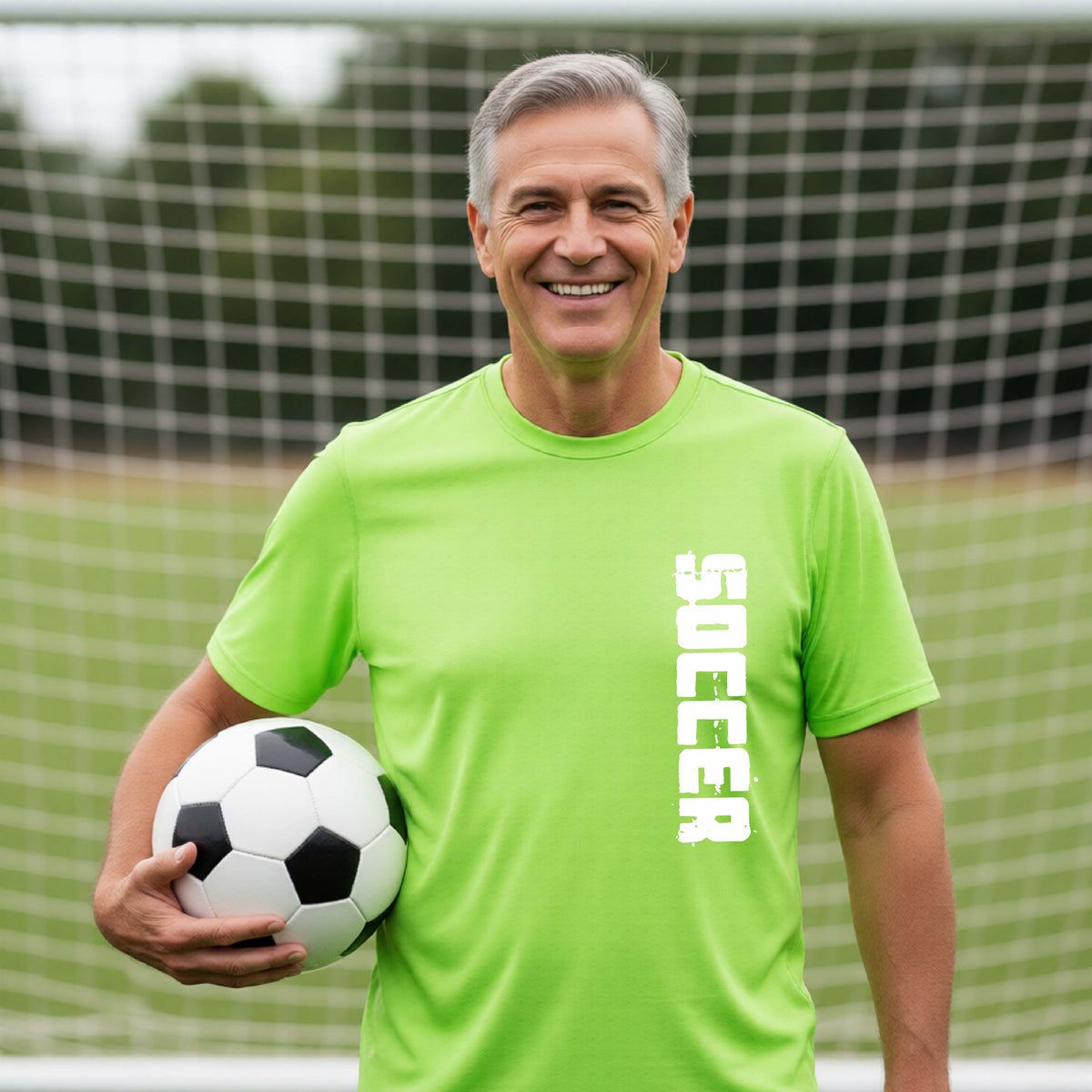 A smiling middle-aged man wearing a bright lime green athletic t-shirt with the word 'SOCCER' printed vertically in white on the right side of the chest, holding a black and white soccer ball in front of a soccer goal net.
