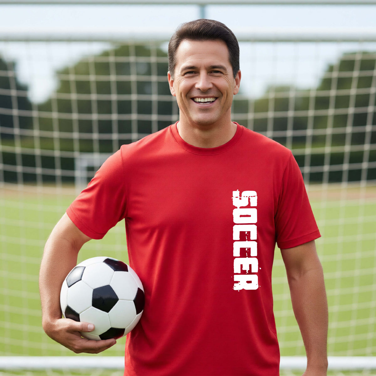 A smiling middle-aged man wearing a red athletic t-shirt with the word 'SOCCER' printed vertically in white on the right side of the chest, holding a black and white soccer ball in front of a soccer goal net.