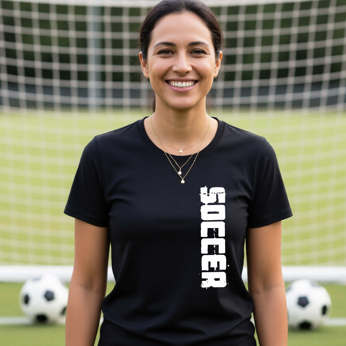 A smiling woman wearing a black short-sleeve t-shirt with the word 'SOCCER' printed vertically in white on the front, standing on a soccer field with a soccer ball in the background.