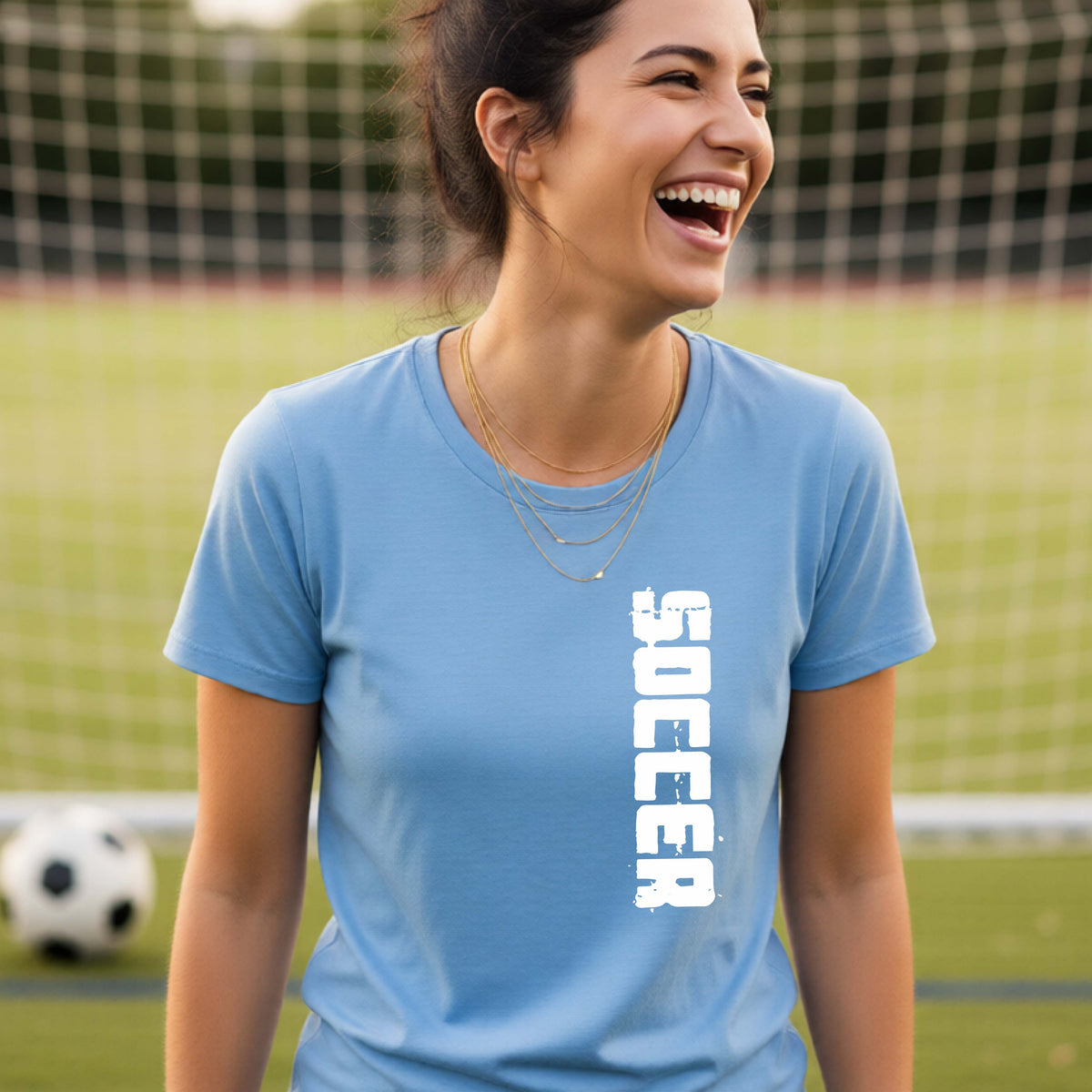 A woman laughing on a soccer field wearing a light blue t-shirt with the word 'SOCCER' printed vertically in white on the front.