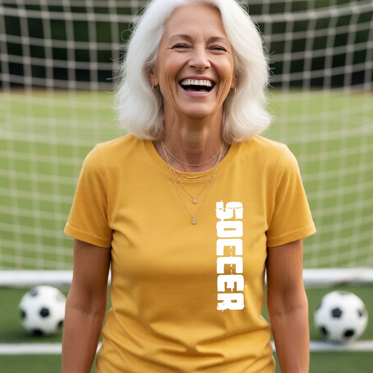 A smiling woman wearing a gold short-sleeve t-shirt with the word 'SOCCER' printed vertically in white on the front, standing on a soccer field with a soccer ball in the background.