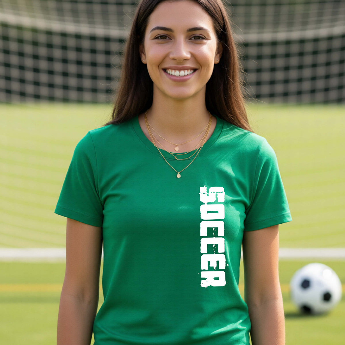 A smiling woman wearing a green short-sleeve t-shirt with the word 'SOCCER' printed vertically in white on the front, standing on a soccer field with a soccer ball in the background.