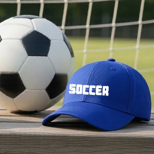 A royal blue baseball cap with 'SOCCER' in white across the front, placed on a wooden surface next to a black and white soccer ball in front of a soccer net.