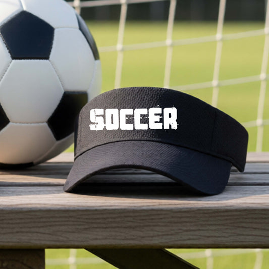 A black visor with the word 'SOCCER' printed in white across the front, resting on a wooden picnic table with a soccer ball and a soccer field in the background.