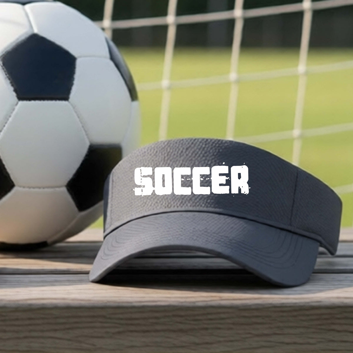 A dark gray visor with the word 'SOCCER' printed in white across the front, resting on a wooden surface with a soccer ball and a blurred soccer field in the background.