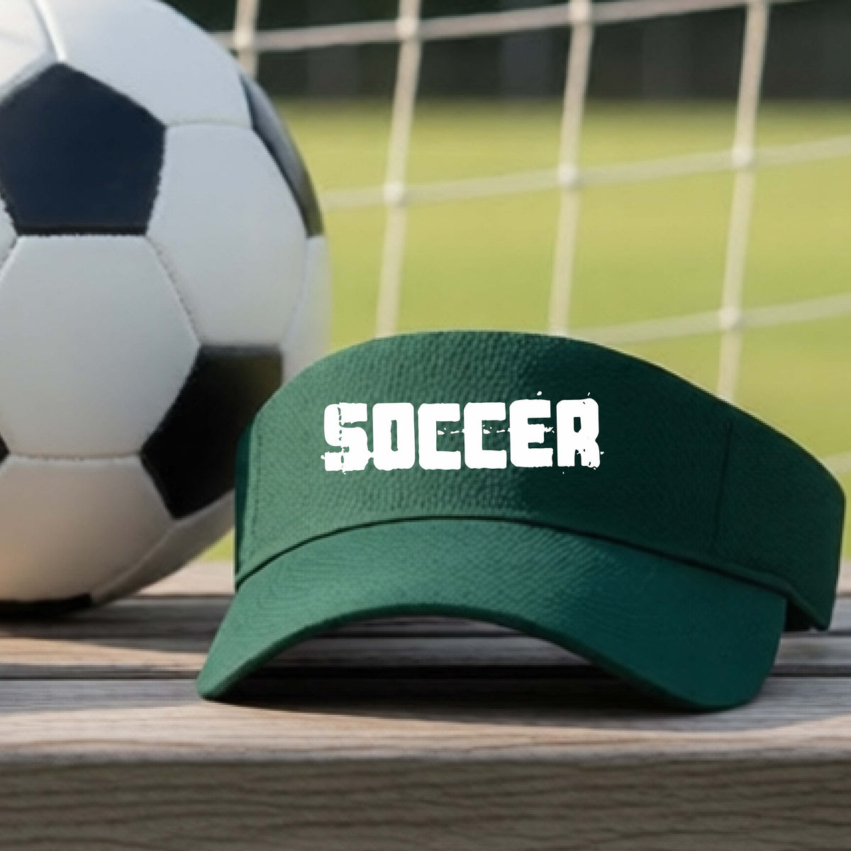 A dark green visor with the word 'SOCCER' printed in white distressed letters, resting on a wooden surface with a soccer ball and a goal net in the background.