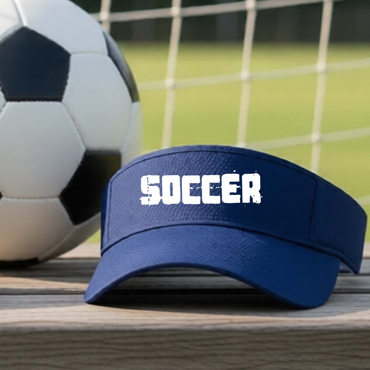 A navy blue visor with the word 'SOCCER' printed in white across the front, resting on a wooden surface with a soccer ball and a blurred soccer field in the background.