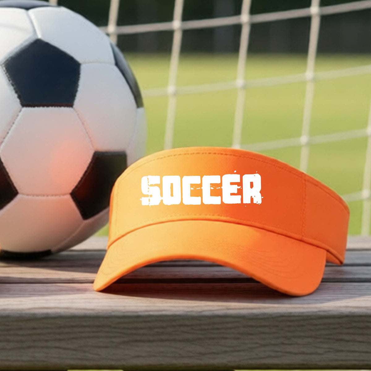 An orange visor with the word 'SOCCER' printed in white across the front, resting on a wooden surface with a soccer ball and a blurred soccer field in the background.