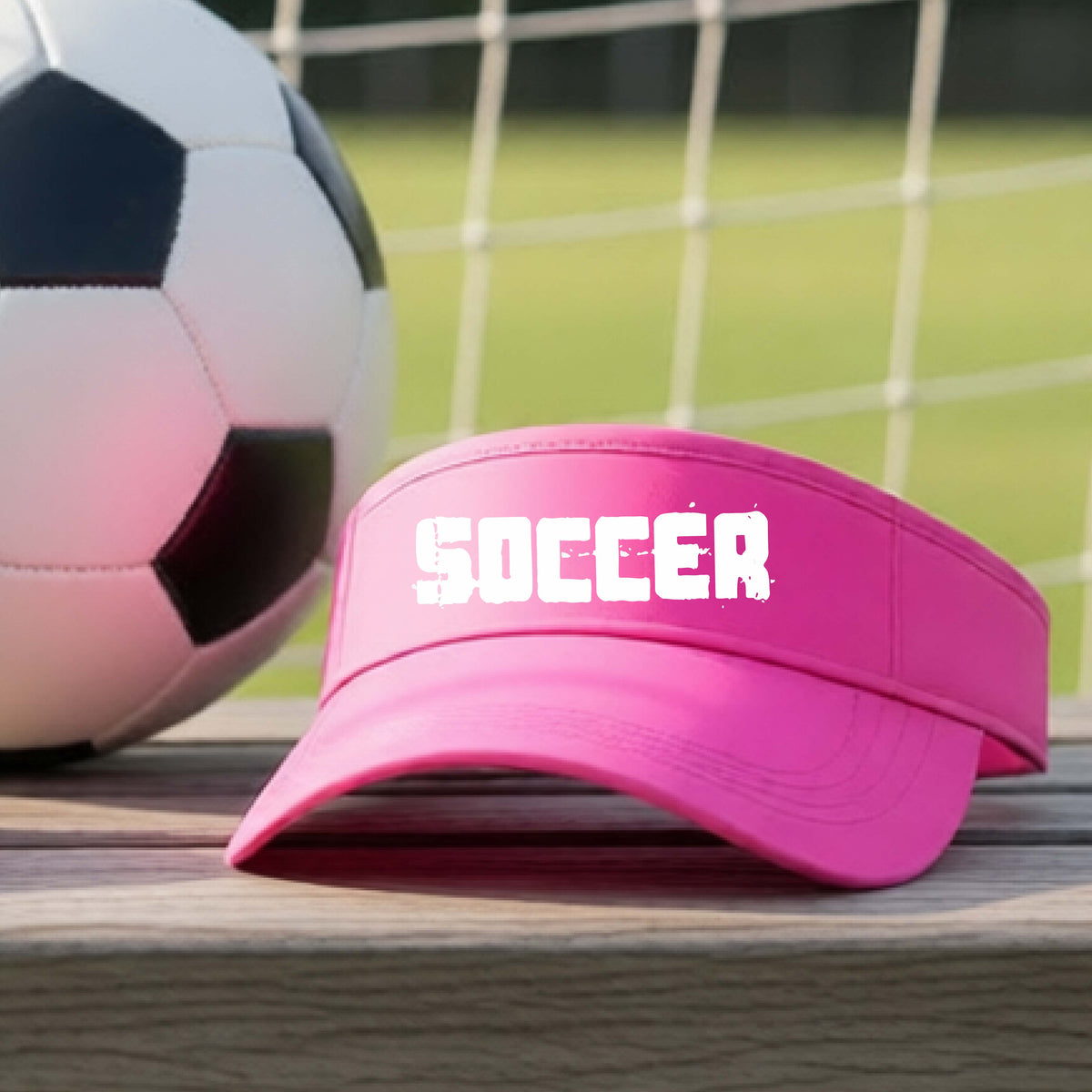 A bright pink visor with the word 'SOCCER' printed in white across the front, resting on a wooden surface with a soccer ball and a blurred soccer field in the background.