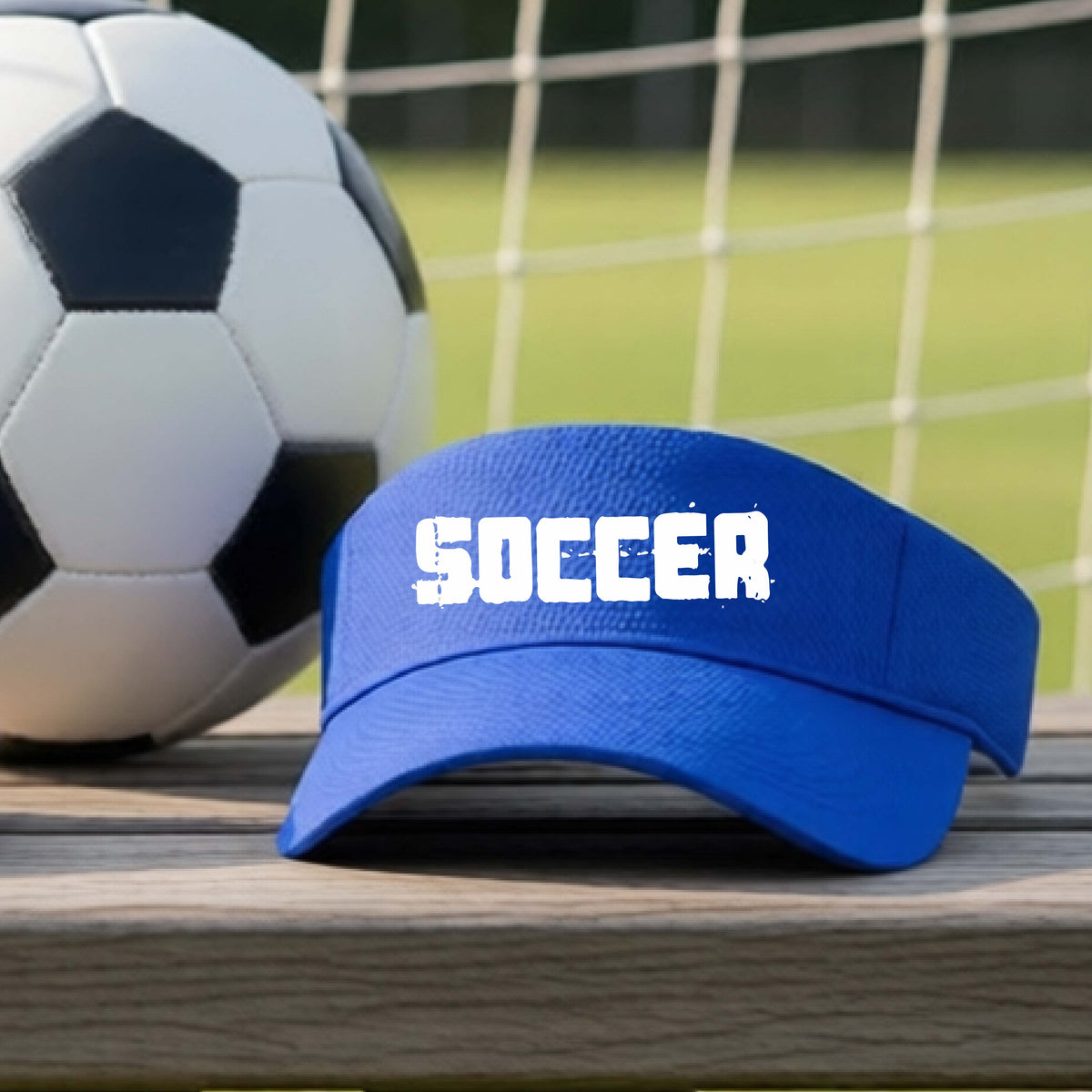 A royal blue visor with the word 'SOCCER' printed in white across the front, resting on a wooden surface with a soccer ball and a soccer net in the background.