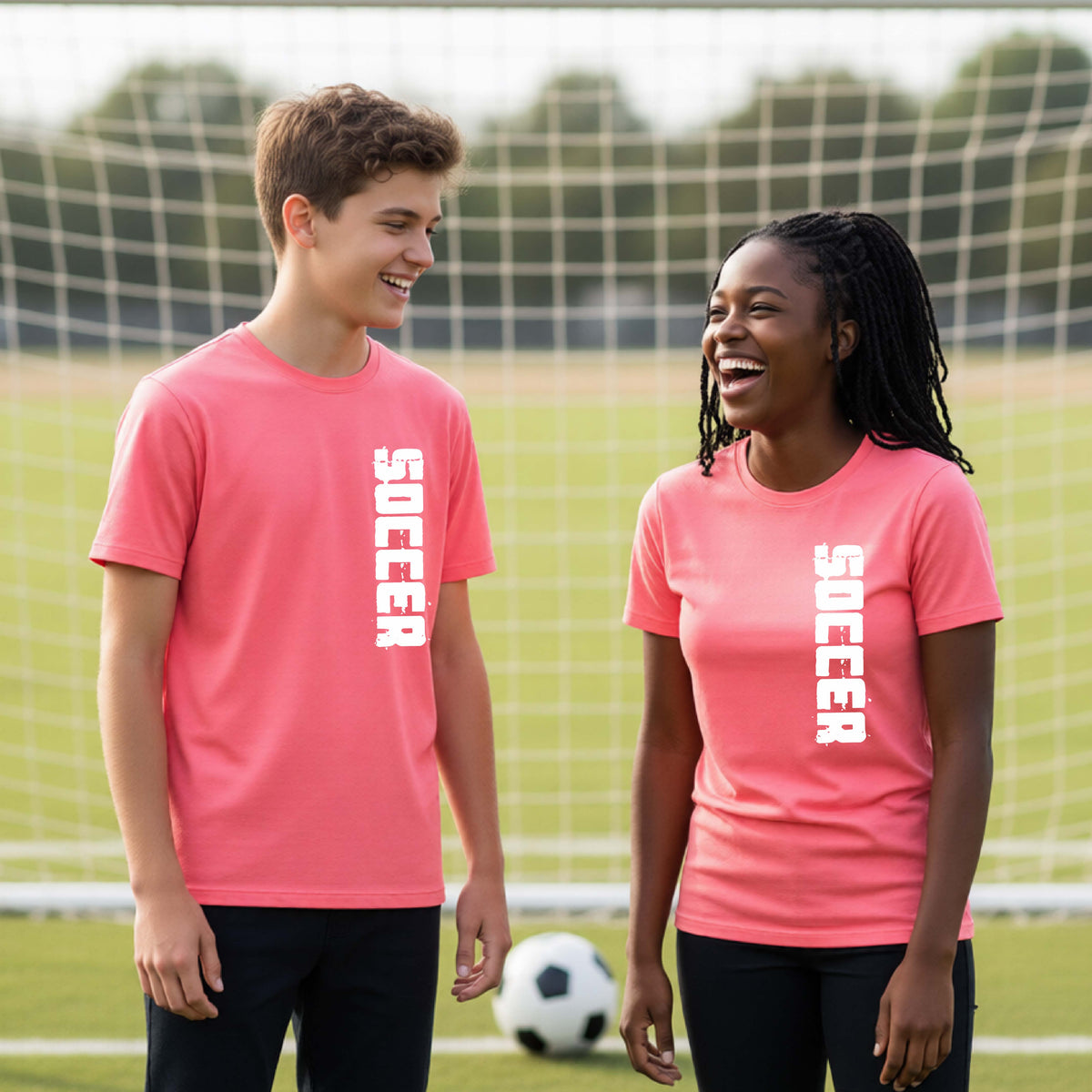 Two young students on a soccer field holding a soccer ball, wearing a oral pink t-shirt with a white graphic of the word 'SOCCER' in block font printed down the side of the shirt