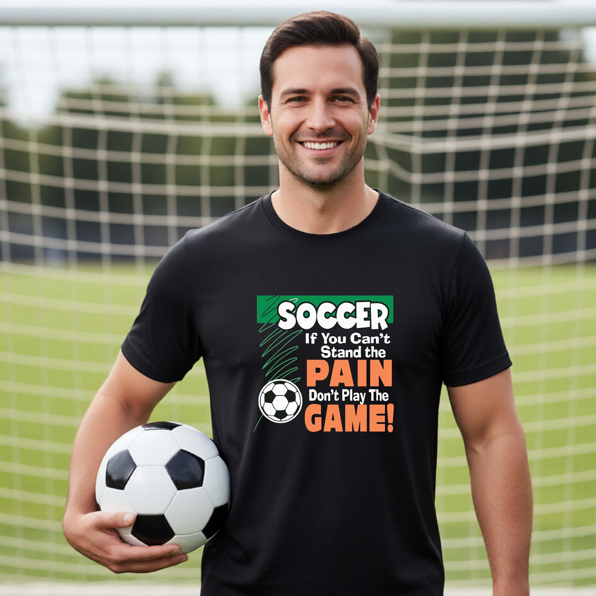 A smiling man on a soccer field holds a black-and-white soccer ball while wearing a black graphic T-shirt with the slogan 'SOCCER If You Can't Stand the PAIN Don't Play The GAME!' in green, white, and orange text.