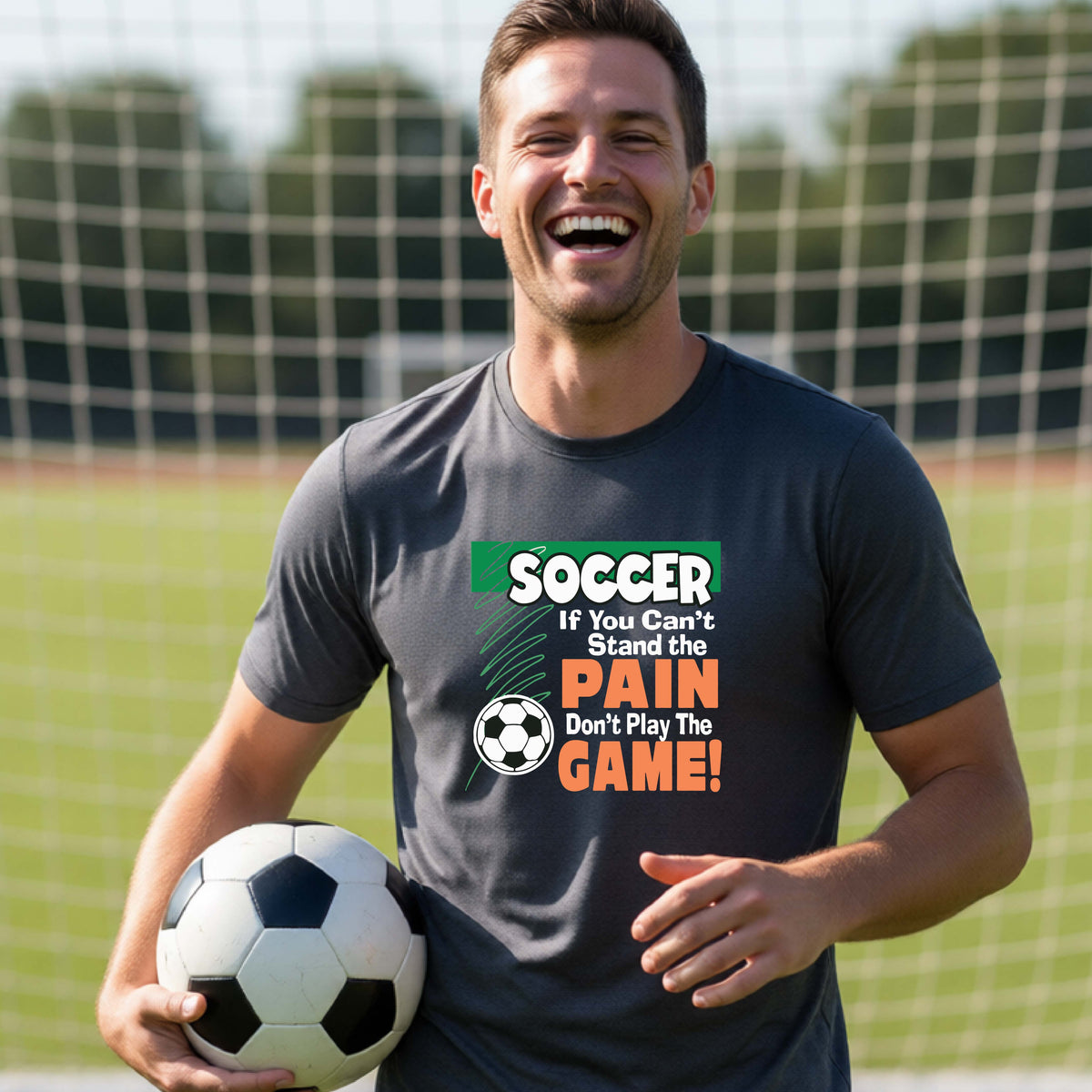 A smiling man on a soccer field holds a black-and-white soccer ball while wearing a dark gray graphic T-shirt with the slogan 'SOCCER If You Can't Stand the PAIN Don't Play The GAME!' in green, white, and orange text.
