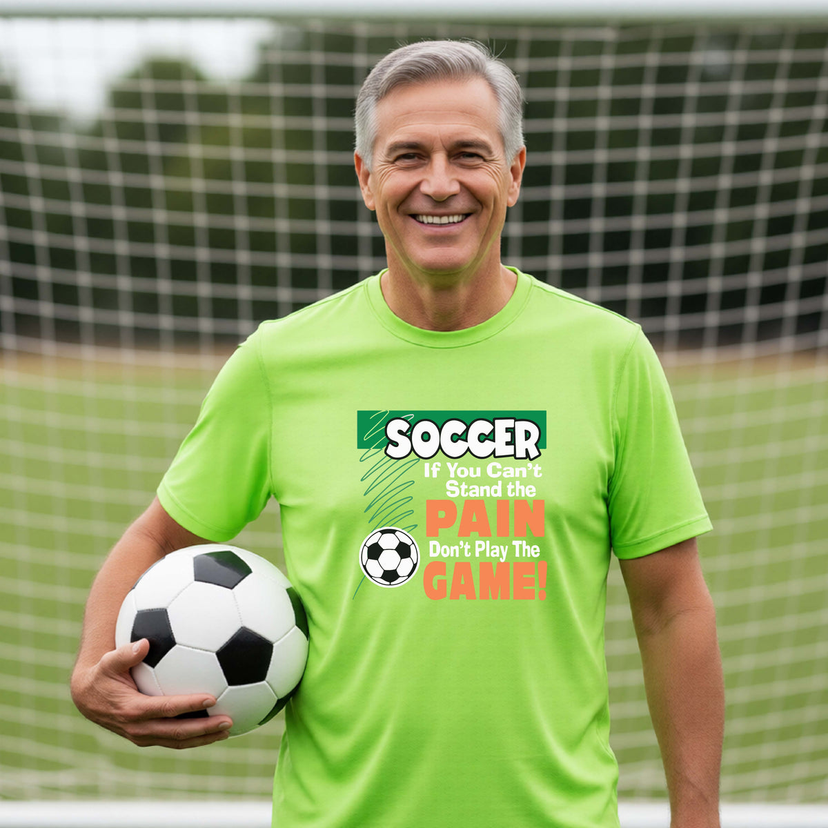 A smiling man wearing a bright lime green graphic t-shirt with a soccer slogan stands on a soccer field holding a black and white soccer ball. The shirt features a green and orange graphic that reads 'SOCCER If You Can't Stand the PAIN Don't Play The GAME!'.