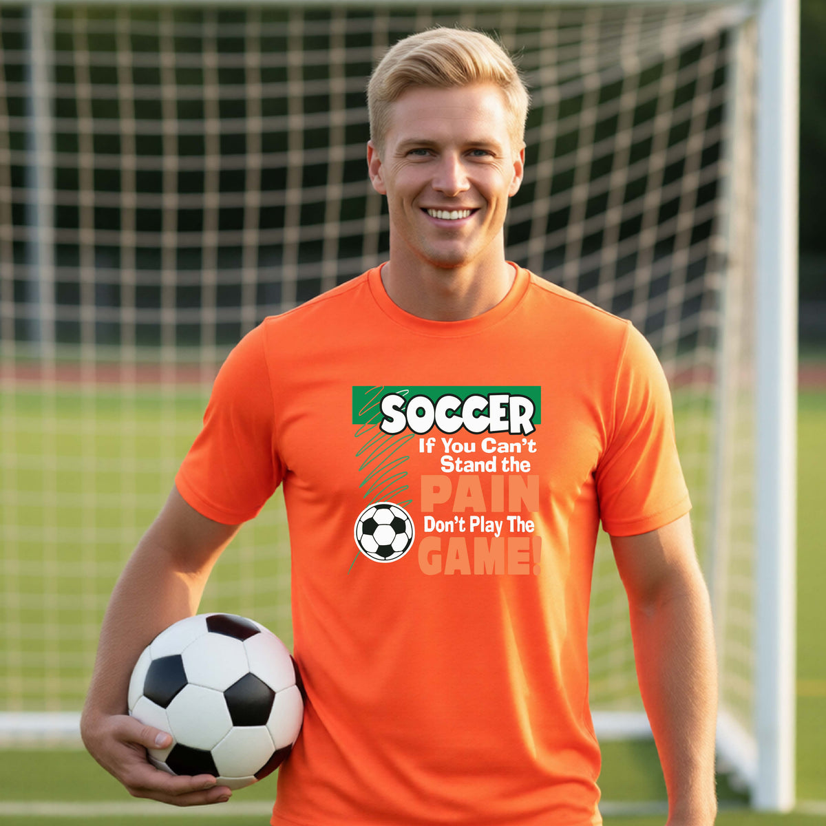 A smiling man on a soccer field holds a black-and-white soccer ball while wearing a bright orange graphic T-shirt with the slogan 'SOCCER If You Can't Stand the PAIN Don't Play The GAME!' in green, white, and orange text.