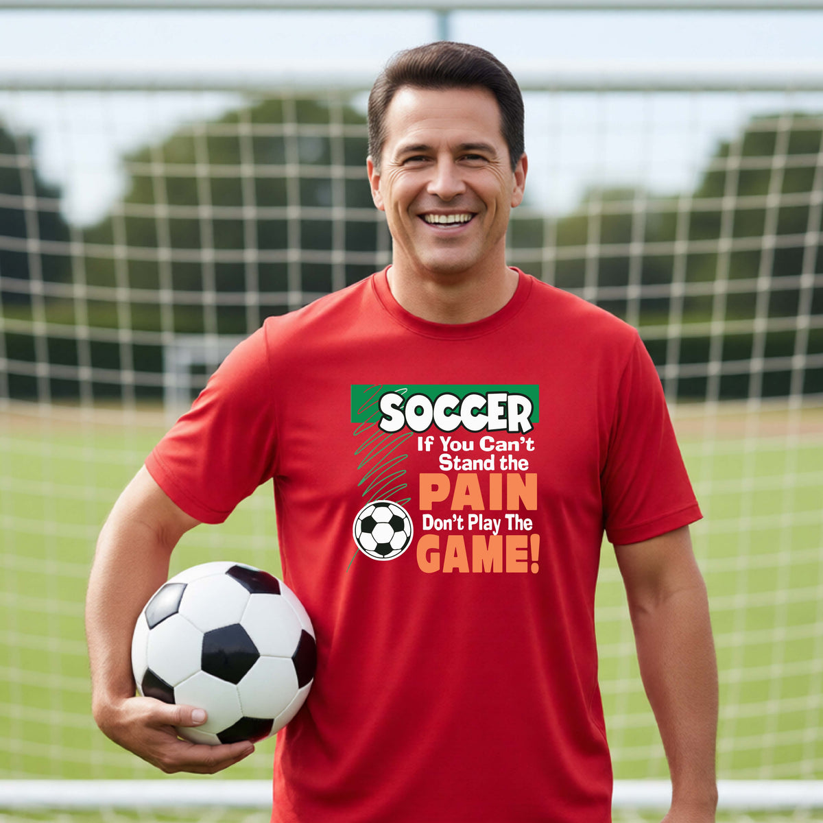 A smiling man on a soccer field holds a black and white soccer ball while wearing a red graphic t-shirt with the text 'SOCCER IF YOU Can't Stand the PAIN Don't Play The GAME!' in green, white, and orange.