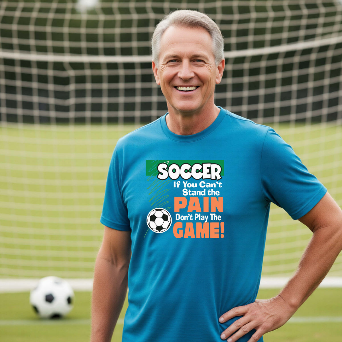 A smiling man wearing a bright blue graphic t-shirt on a soccer field with a goal net in the background. The shirt features a green and orange graphic that reads 'SOCCER IF YOU Can't Stand the PAIN Don't Play The GAME!' with a soccer ball icon.