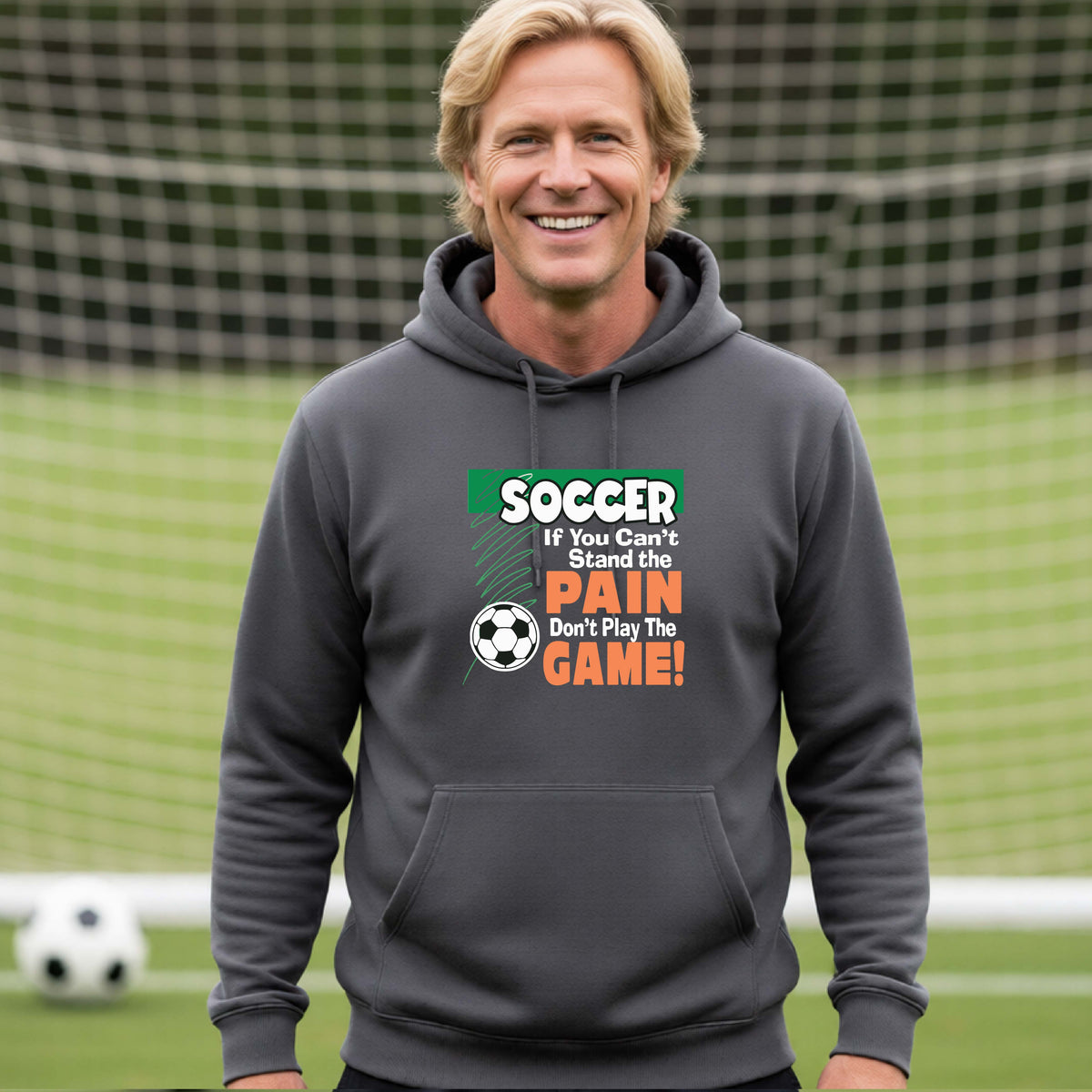 A smiling man on a soccer field while wearing a dark gray hoodie pullover with the graphic slogan 'SOCCER If You Can't Stand the PAIN Don't Play The GAME!' in green, white, and orange text.