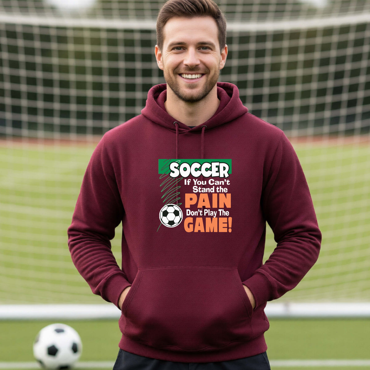 A smiling man on a soccer field while wearing a maroon hoodie pullover with the graphic slogan 'SOCCER If You Can't Stand the PAIN Don't Play The GAME!' in green, white, and orange text.