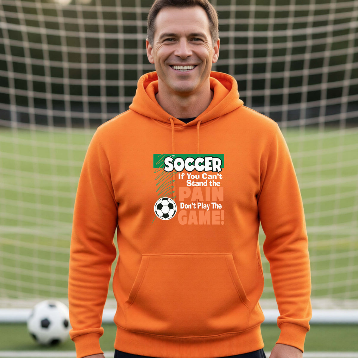 A smiling man on a soccer field while wearing an orange hoodie pullover with the graphic slogan 'SOCCER If You Can't Stand the PAIN Don't Play The GAME!' in green, white, and orange text.