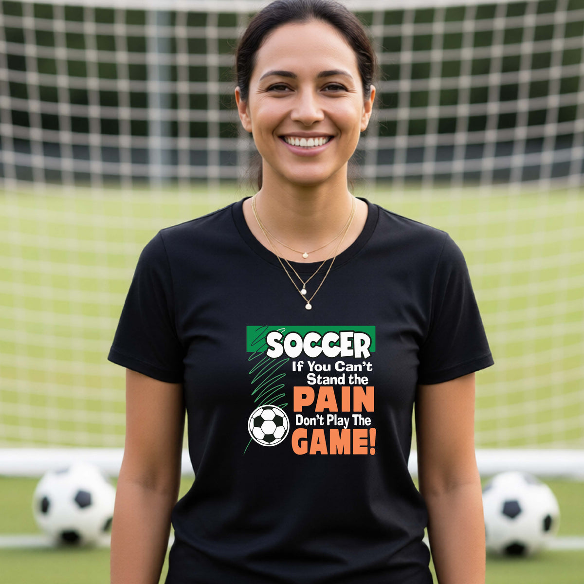 A smiling woman on a soccer field while wearing a black graphic T-shirt with the slogan 'SOCCER If You Can't Stand the PAIN Don't Play The GAME!' in green, white, and orange text.