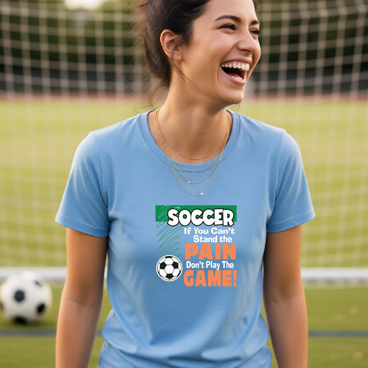 A smiling woman on a soccer field while wearing a blue graphic T-shirt with the slogan 'SOCCER If You Can't Stand the PAIN Don't Play The GAME!' in green, white, and orange text.