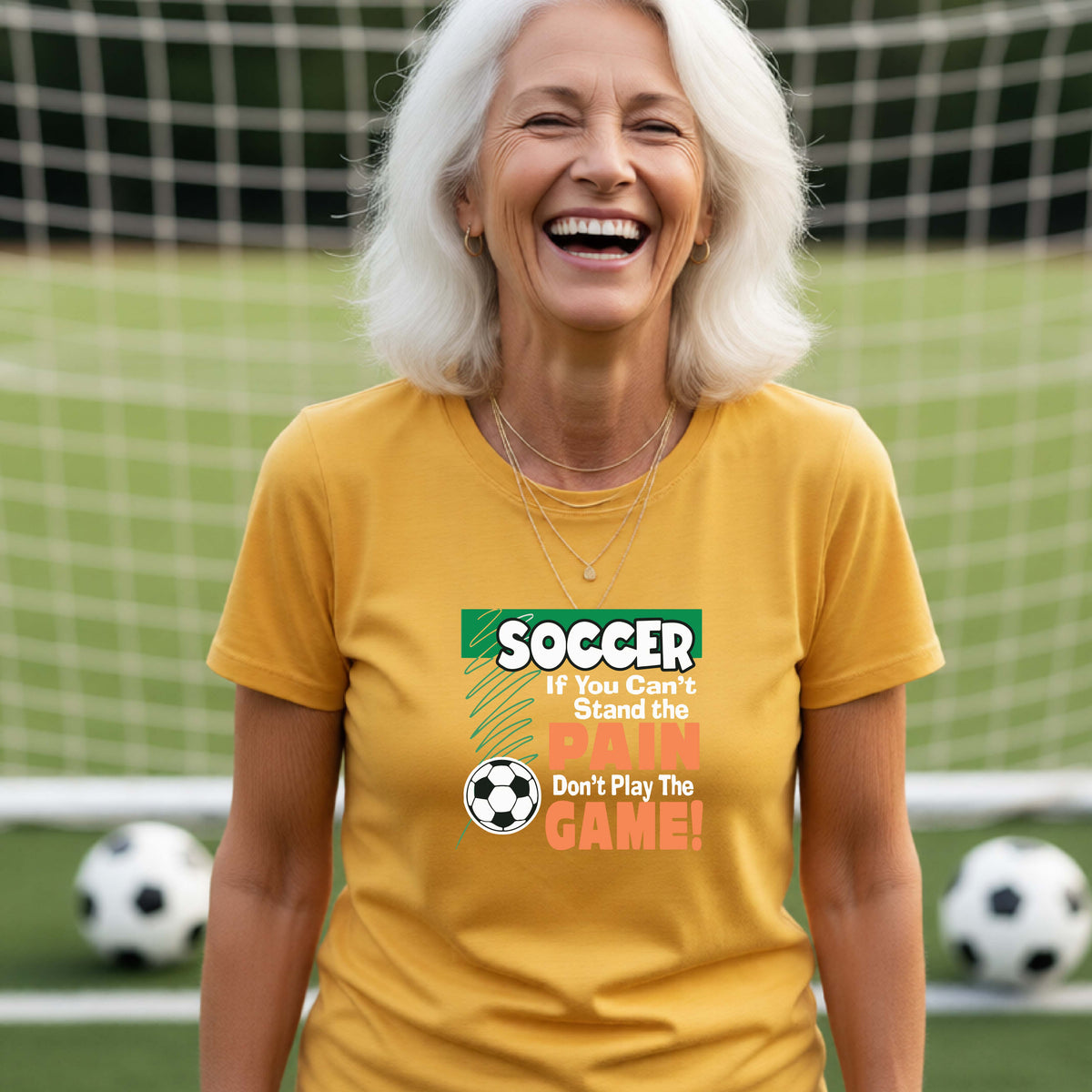 A smiling woman on a soccer field while wearing a gold graphic T-shirt with the slogan 'SOCCER If You Can't Stand the PAIN Don't Play The GAME!' in green, white, and orange text.