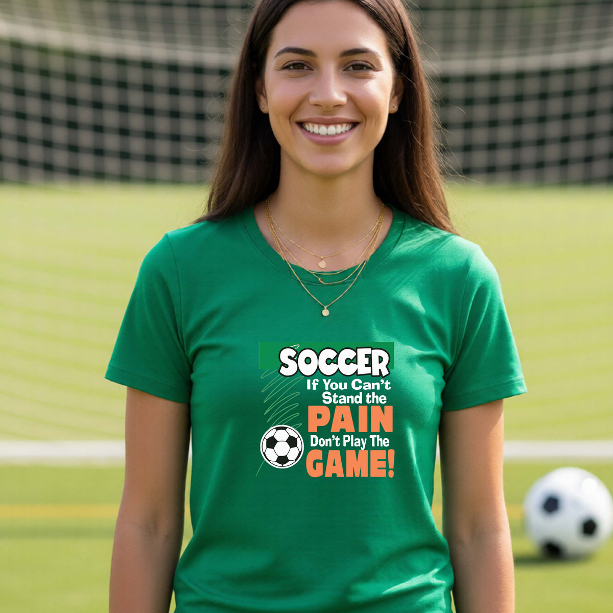 A smiling woman on a soccer field while wearing a green graphic T-shirt with the slogan 'SOCCER If You Can't Stand the PAIN Don't Play The GAME!' in green, white, and orange text.