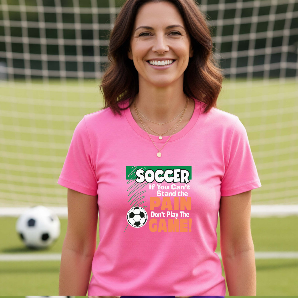 A smiling woman on a soccer field while wearing a pink graphic T-shirt with the slogan 'SOCCER If You Can't Stand the PAIN Don't Play The GAME!' in green, white, and orange text.