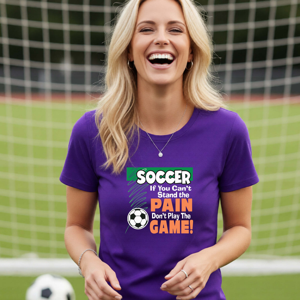 A smiling woman on a soccer field while wearing a purple graphic T-shirt with the slogan 'SOCCER If You Can't Stand the PAIN Don't Play The GAME!' in green, white, and orange text.