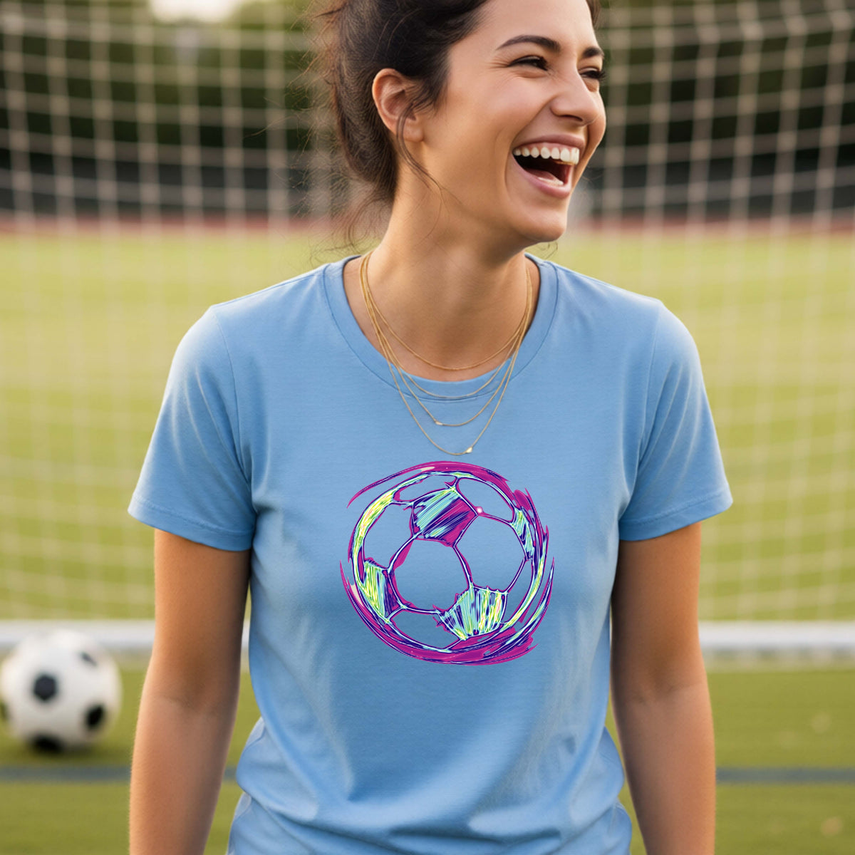 A woman smiling on a soccer field wearing a blue t-shirt with a colorful, iridescent soccer ball graphic on the chest.