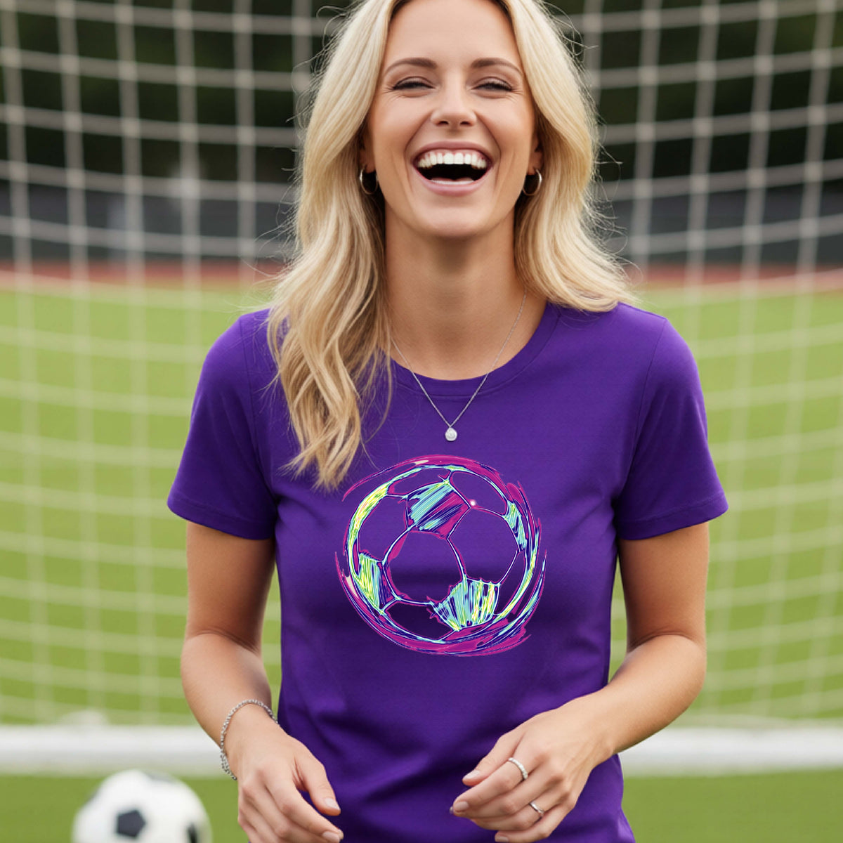 A woman with blonde hair smiling and laughing, wearing a purple short-sleeve t-shirt with a colorful soccer ball graphic on the front, standing in front of a soccer goal.
