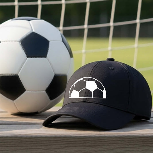 A black baseball cap with a white soccer ball graphic on the front, resting on a wooden surface in front of a soccer ball and goal net on a grassy field.