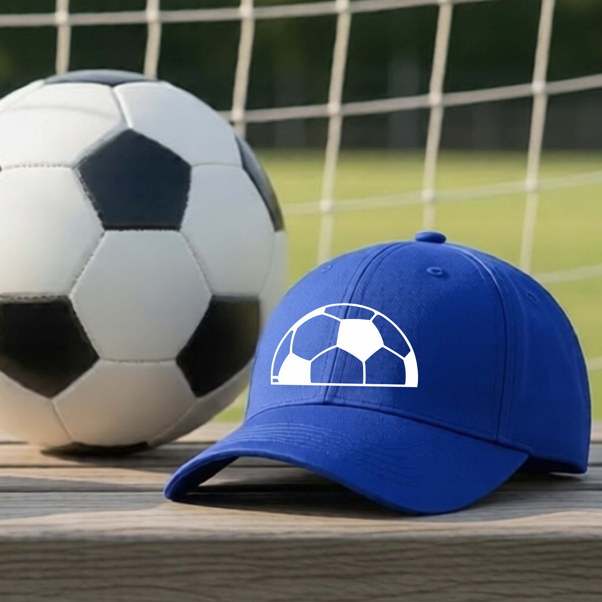 A royal blue baseball cap with a white soccer ball graphic on the front, resting on a wooden surface in front of a soccer ball and goal net.