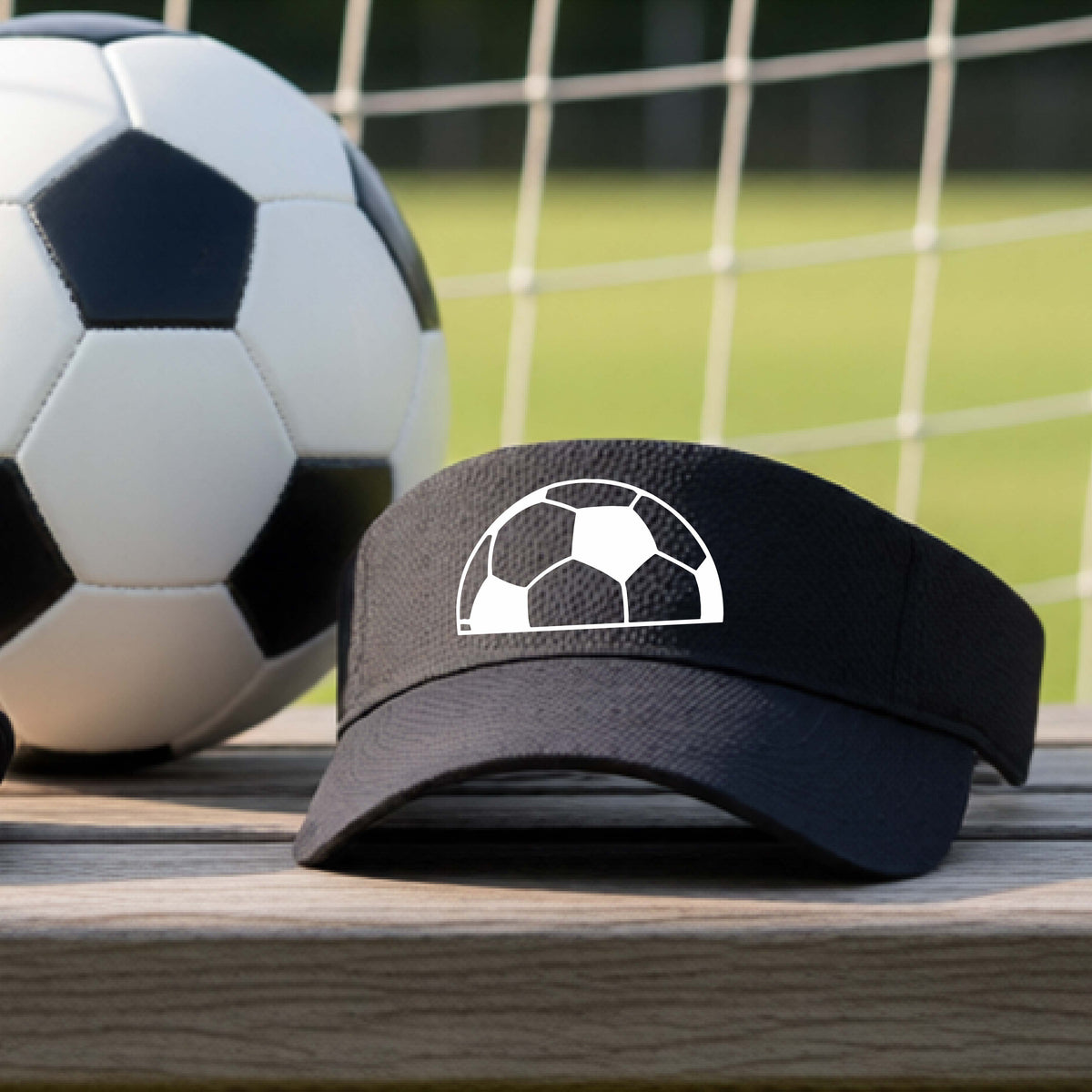 A black baseball cap with a white soccer ball graphic on the front, resting on a wooden surface with a soccer ball and a goal net in the blurred background.