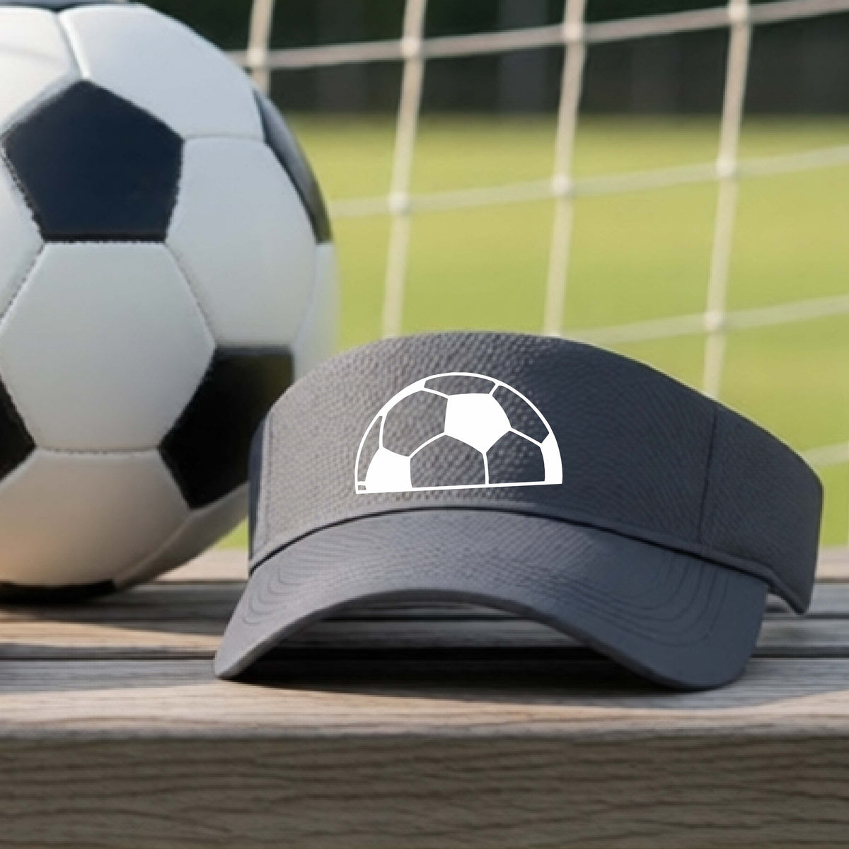 A gray baseball cap with a white soccer ball graphic on the front, resting on a wooden surface with a soccer ball and a goal net in the blurred background.
