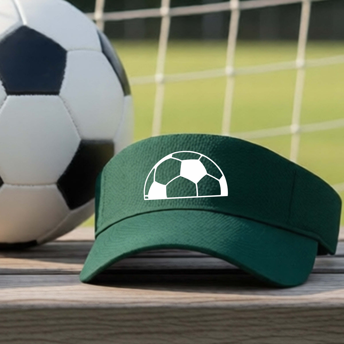 A green baseball cap with a white soccer ball graphic on the front, resting on a wooden surface with a soccer ball and a goal net in the blurred background.