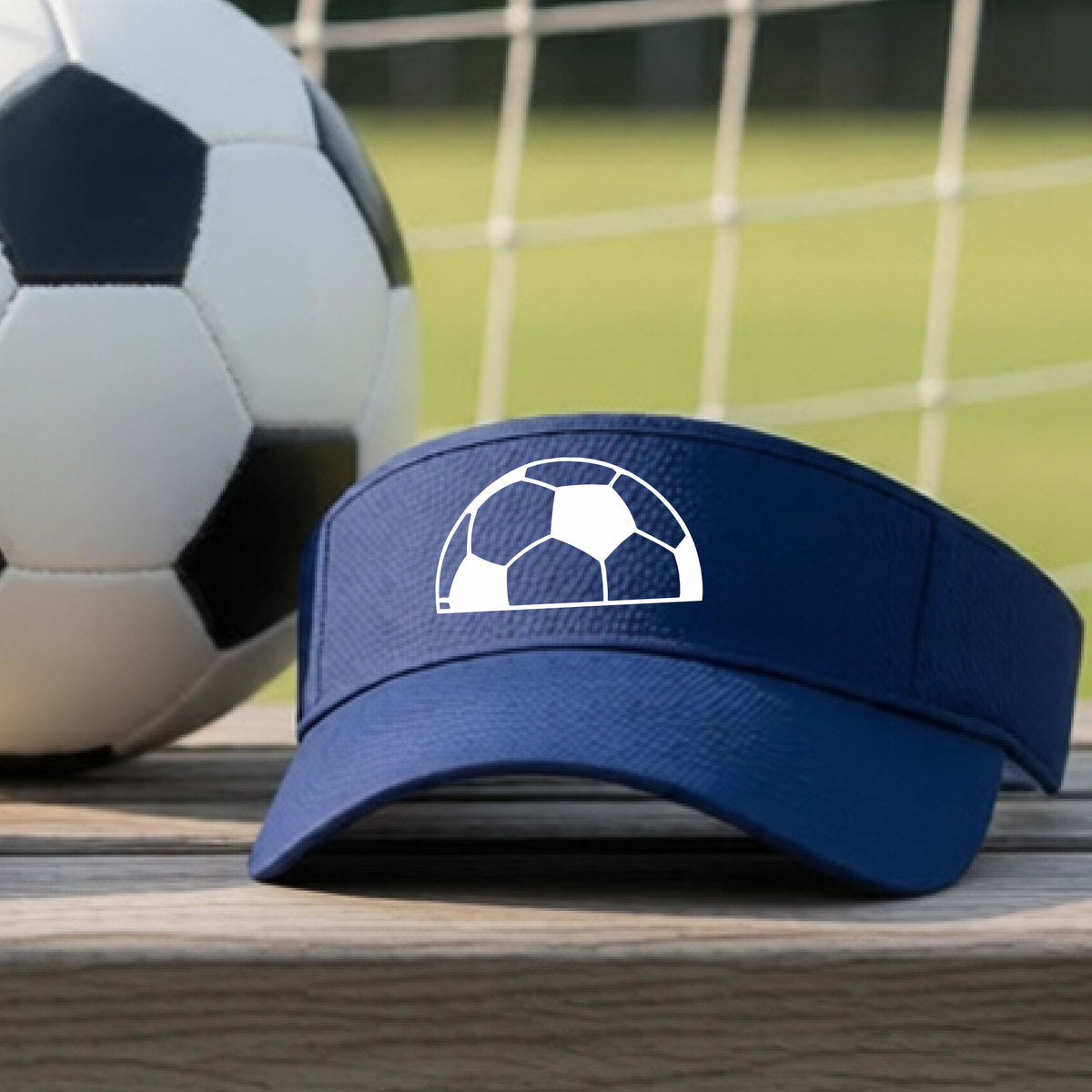 A navy blue baseball cap with a white soccer ball graphic on the front, resting on a wooden surface with a soccer ball and a goal net in the blurred background.