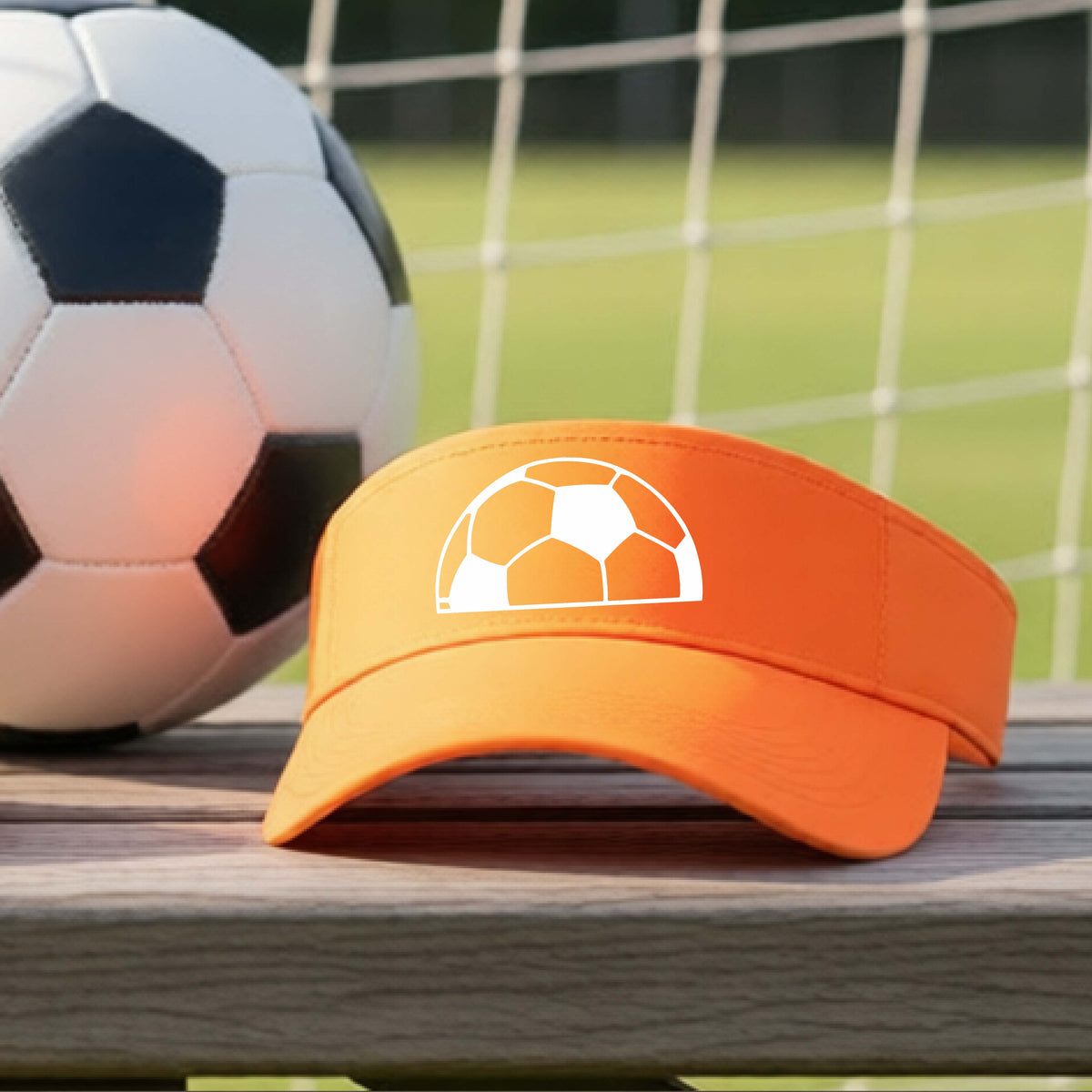 An orange baseball cap with a white soccer ball graphic on the front, resting on a wooden surface with a soccer ball and a goal net in the blurred background.