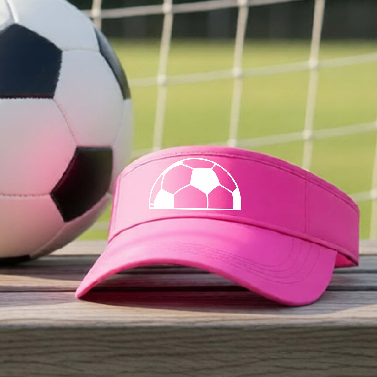 A pink baseball cap with a white soccer ball graphic on the front, resting on a wooden surface with a soccer ball and a goal net in the blurred background.